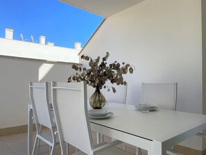 Un espace repas moderne avec une table et des chaises blanches, un vase contenant des feuilles s échées et une vue dégagée sur le ciel grâce à une lucarne.