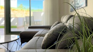 A modern living room with a grey sofa, a round coffee table, and a large window with a view of a garden and a building. - Belvilla by OYO Apartamento Moja!car (Mojacar)