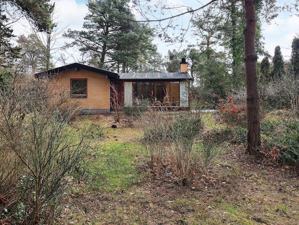A single-story house with a brick exterior and a small porch, surrounded by dense woodland.