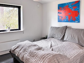 A bedroom with a bed, a nightstand, a wall-mounted painting, and a window with a view of trees.