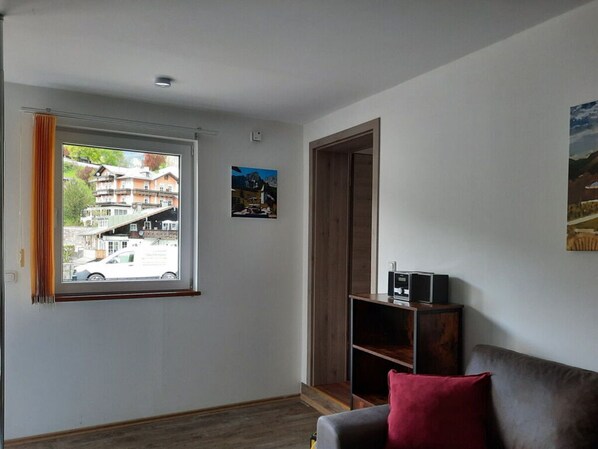 Una habitación con ventana que da a un edificio, una puerta de madera, una pequeña estantería con un dispositivo y un cojín rojo en un sofá.