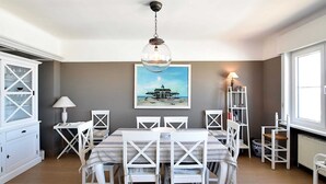 Ein Essbereich mit einem Tisch für sechs Personen, weißen Stühlen, einer Pendelleuchte, einem Gemälde mit Strandmotiv und einem Fenster, das für natürliches Licht sorgt.