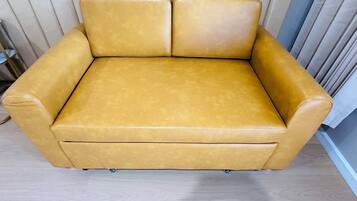 A tan leather sofa with a wooden floor and a curtain in the background.