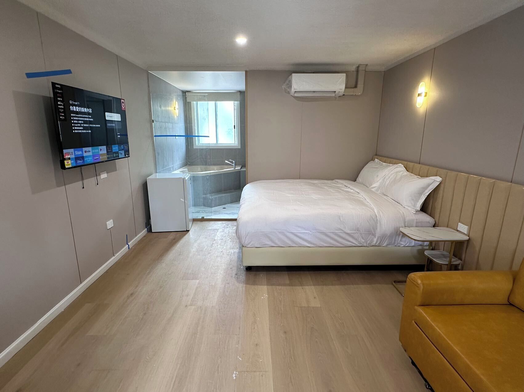 A modern bedroom with a large bed, a small desk, a TV mounted on the wall, and a yellow armchair.