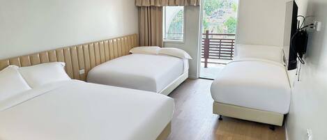Habitación de hotel con tres camas, piso de madera, ventana con vistas a zonas verdes y un televisor montado en la pared.