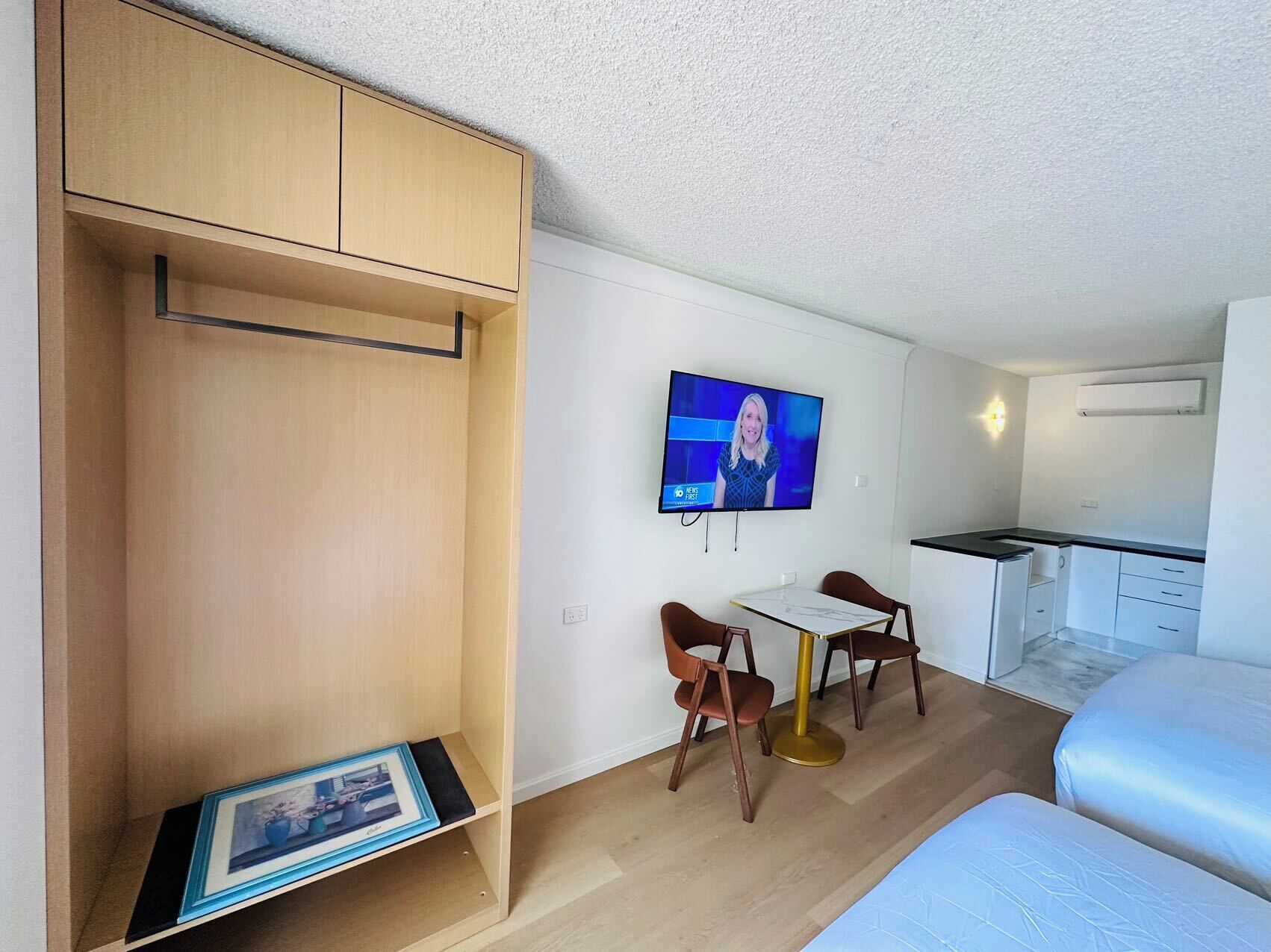 A compact hotel room with a bed, a small kitchenette, a TV mounted on the wall, and a wooden wardrobe.