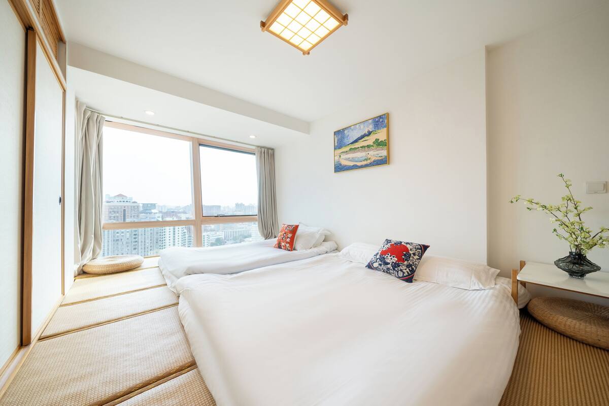 A hotel room with two beds, a large window with a city view, a painting on the wall, and a small table with a vase of flowers.