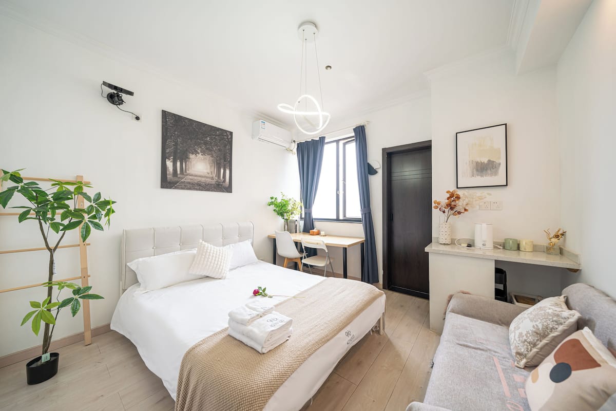 A modern hotel room with a bed, bedside table, desk, and a potted plant.