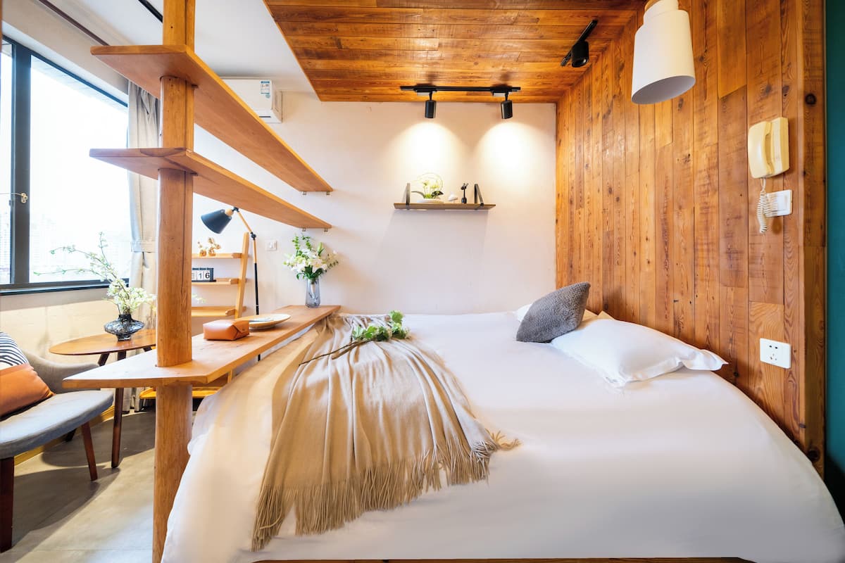A cozy bedroom with a wooden ceiling, a bed with a beige throw, a bedside table, and a small shelf with decorative items.