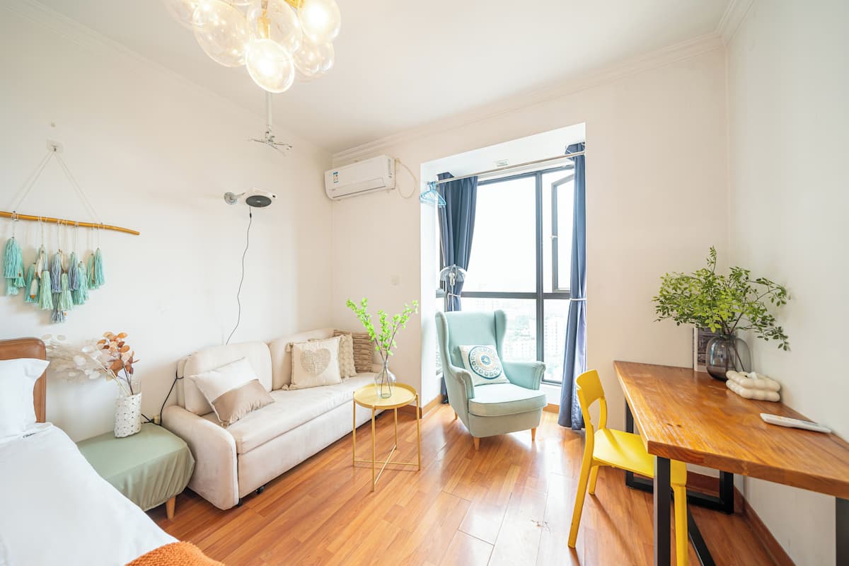 A well-lit living room with a sofa, armchair, and dining table. There is a window with curtains, a hanging plant, and a wall-mounted air conditioner.