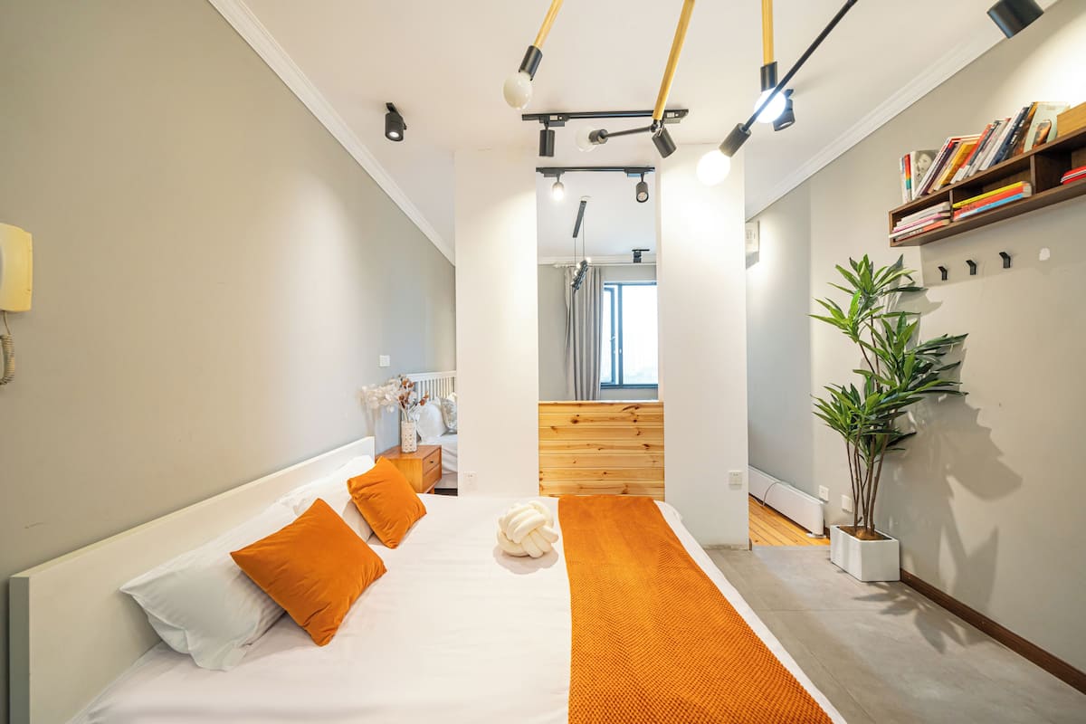 A modern bedroom with a large bed, orange pillows, a white rug, and a potted plant.