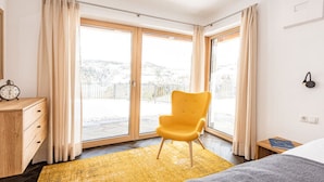 Une chambre à coucher avec un lit, une chaise jaune, une commode en bois et une vue sur un paysage enneigé par de grandes fenêtres.