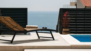 Zona junto a la piscina con una tumbona, una mesa pequeña, una planta en maceta y vistas al mar.