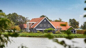 Een groot, donker gebouw met een rood dak, gelegen aan een waterpartij, omgeven door begroeiing en bomen.