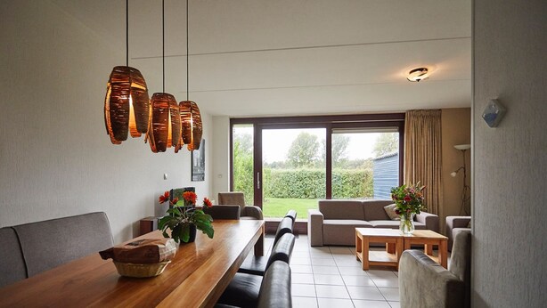 Een eethoek met een houten tafel, banken en hangende hanglampen. Er is een bank en een kleine tafel met een bloemenvaas.