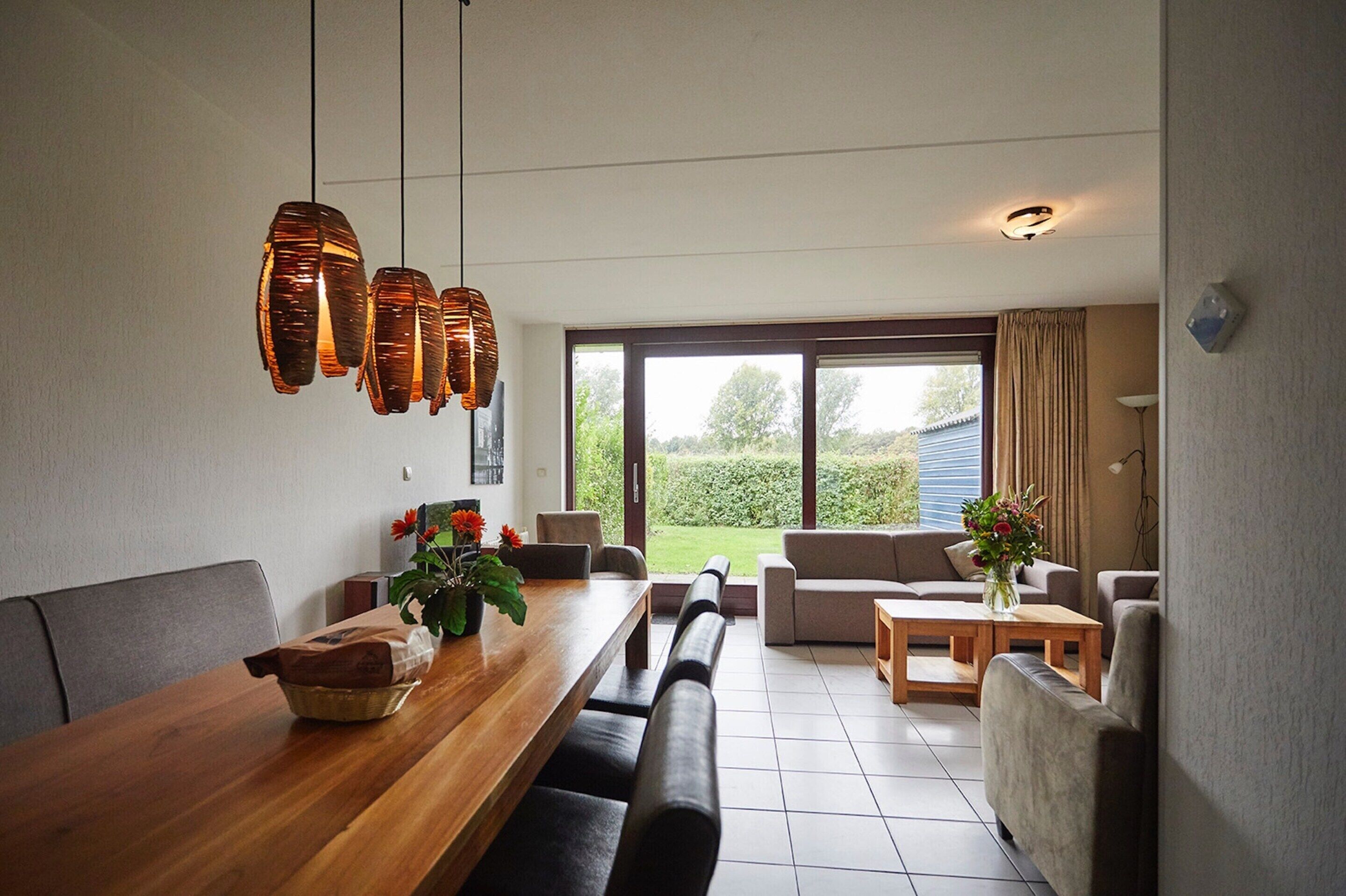 A modern dining area with a wooden table, benches, and hanging pendant lights. There is a sofa and a small coffee table in the background.