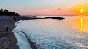 A beach scene with calm waters reflecting the sunset, a pier extending into the water, and a few people walking along the shore. - 5 Person Holiday Home in Melby (Melby)