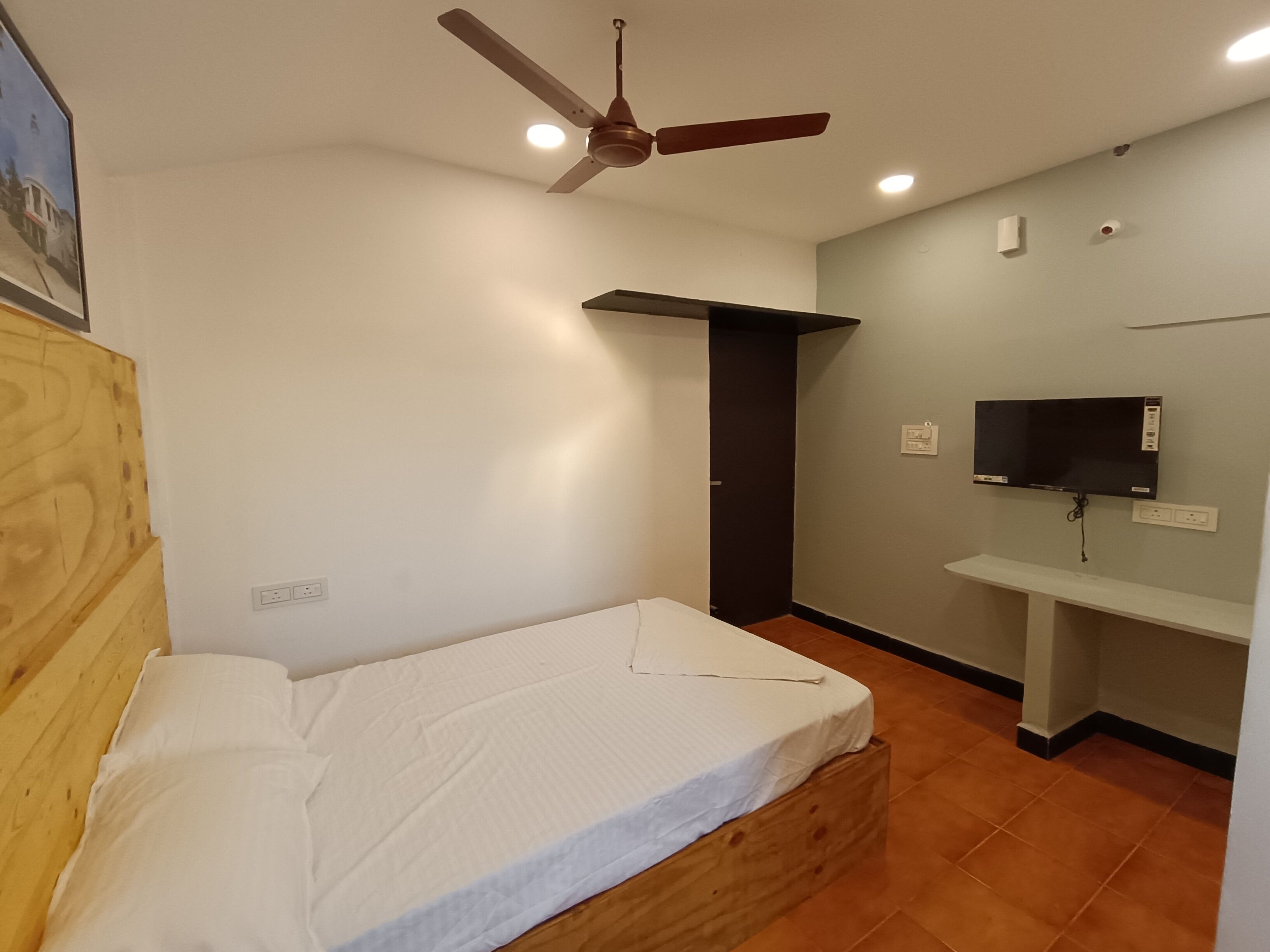 Een kleine, schone kamer met een enkel bed, een plafondventilator, een aan de muur bevestigde televisie en een houten bureau.