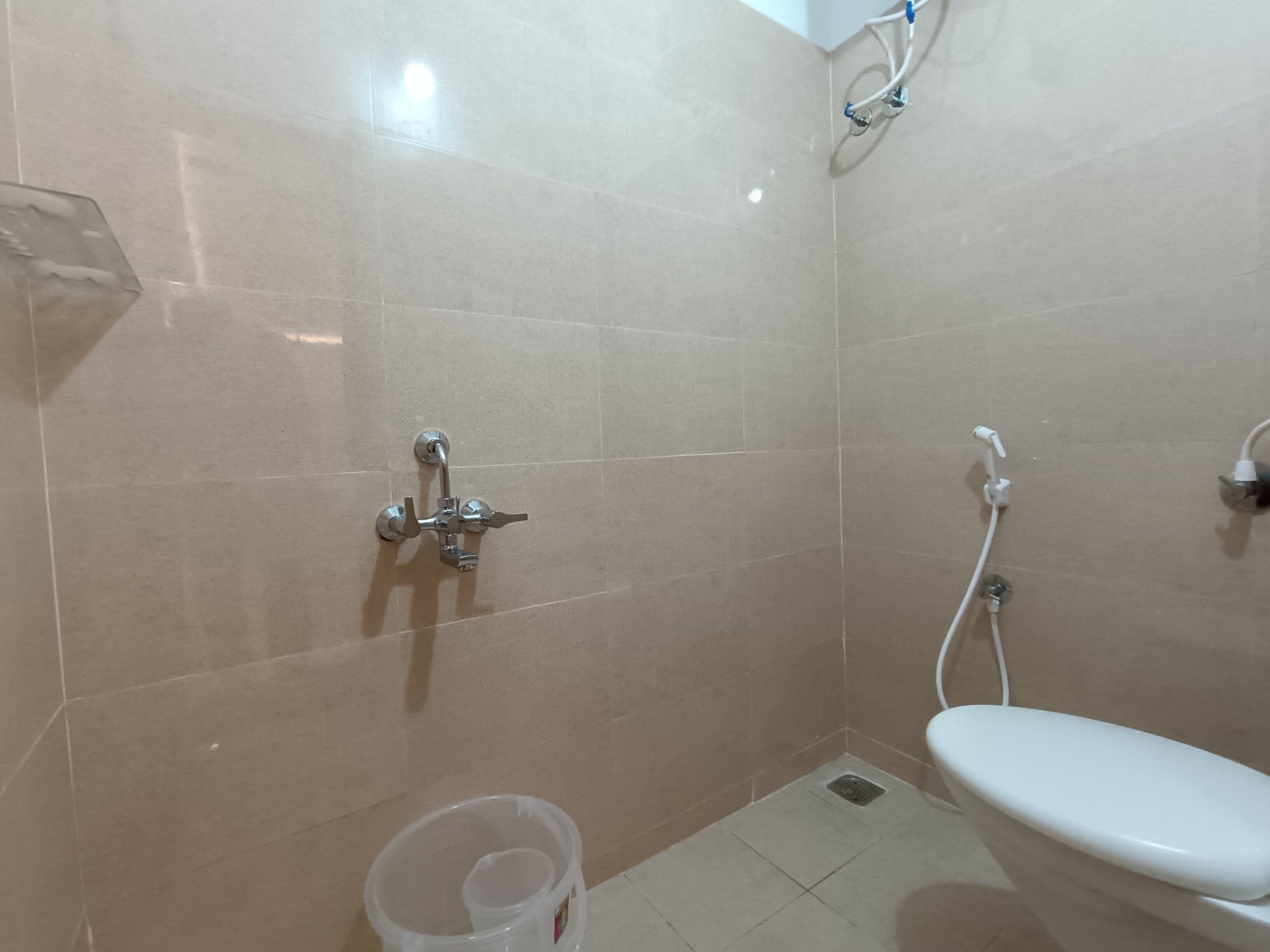 A small, tiled bathroom with a toilet, a shower unit, and a plastic container on the floor.