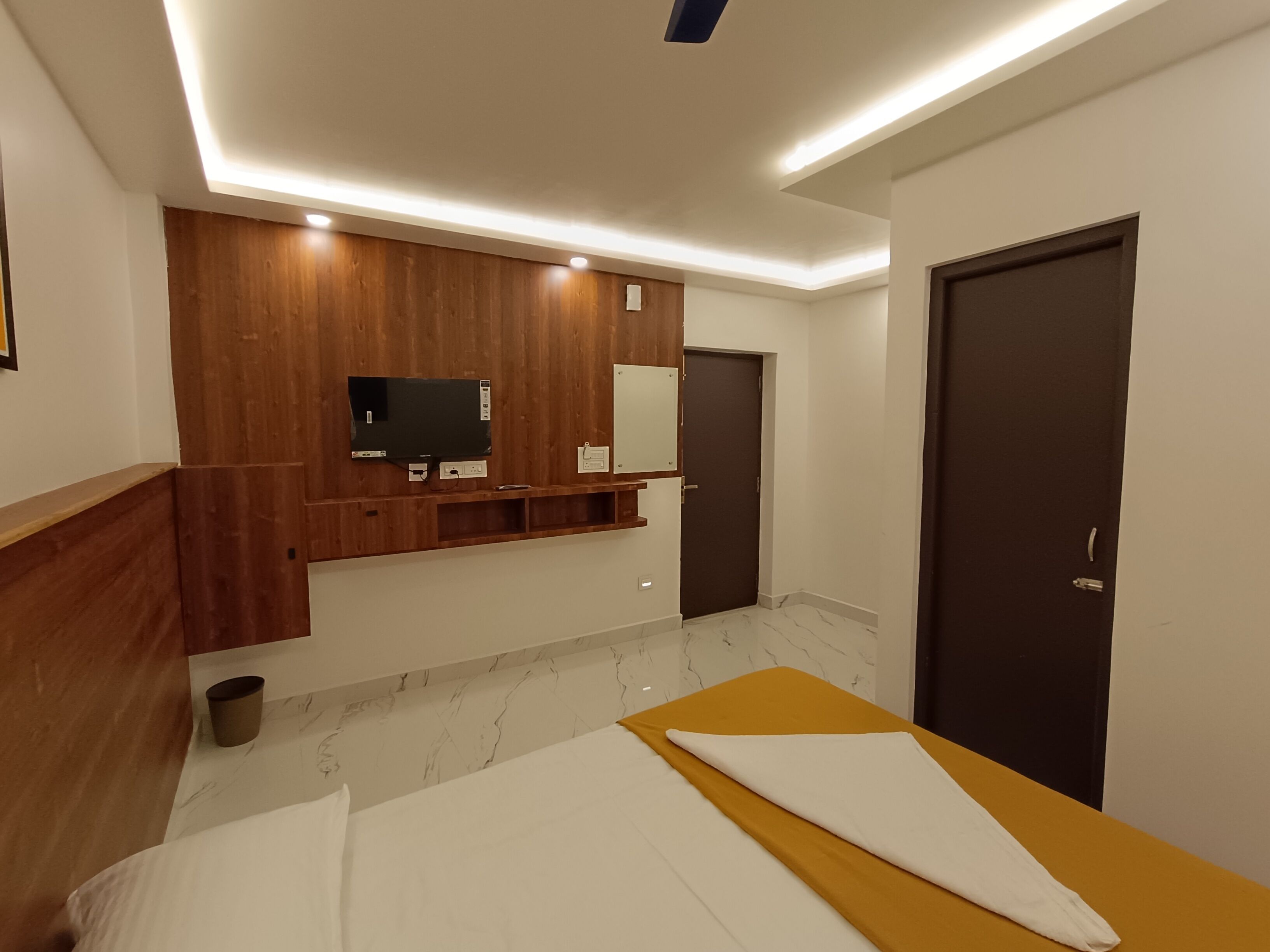 Une chambre d’hôtel moderne avec un mur d’accent en bois, un téléviseur à écran plat, un grand miroir, un lit avec une tête de lit jaune et une porte sombre.