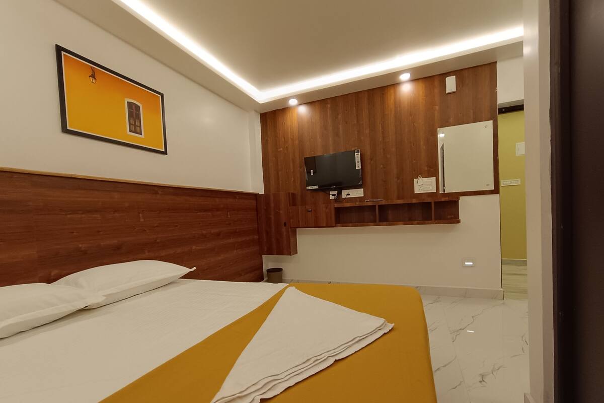 A hotel room with a wooden headboard, a bed with white linens, a yellow bedspread, a flat-screen TV mounted on a wooden panel, and a mirror above the TV.