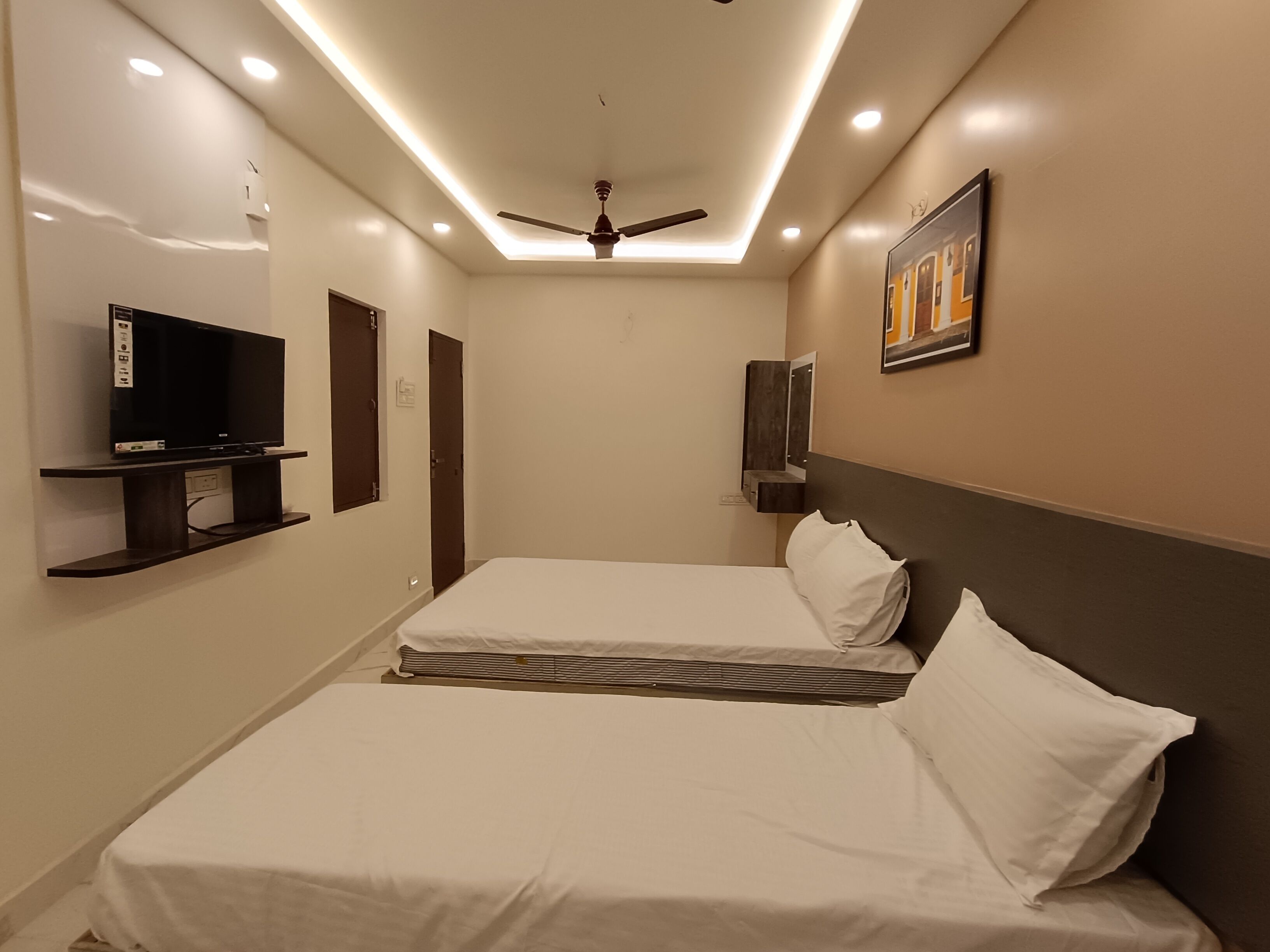 Une chambre d’hôtel avec un lit, un téléviseur posé sur une étagère, un ventilateur de plafond et un tableau accroché au mur.