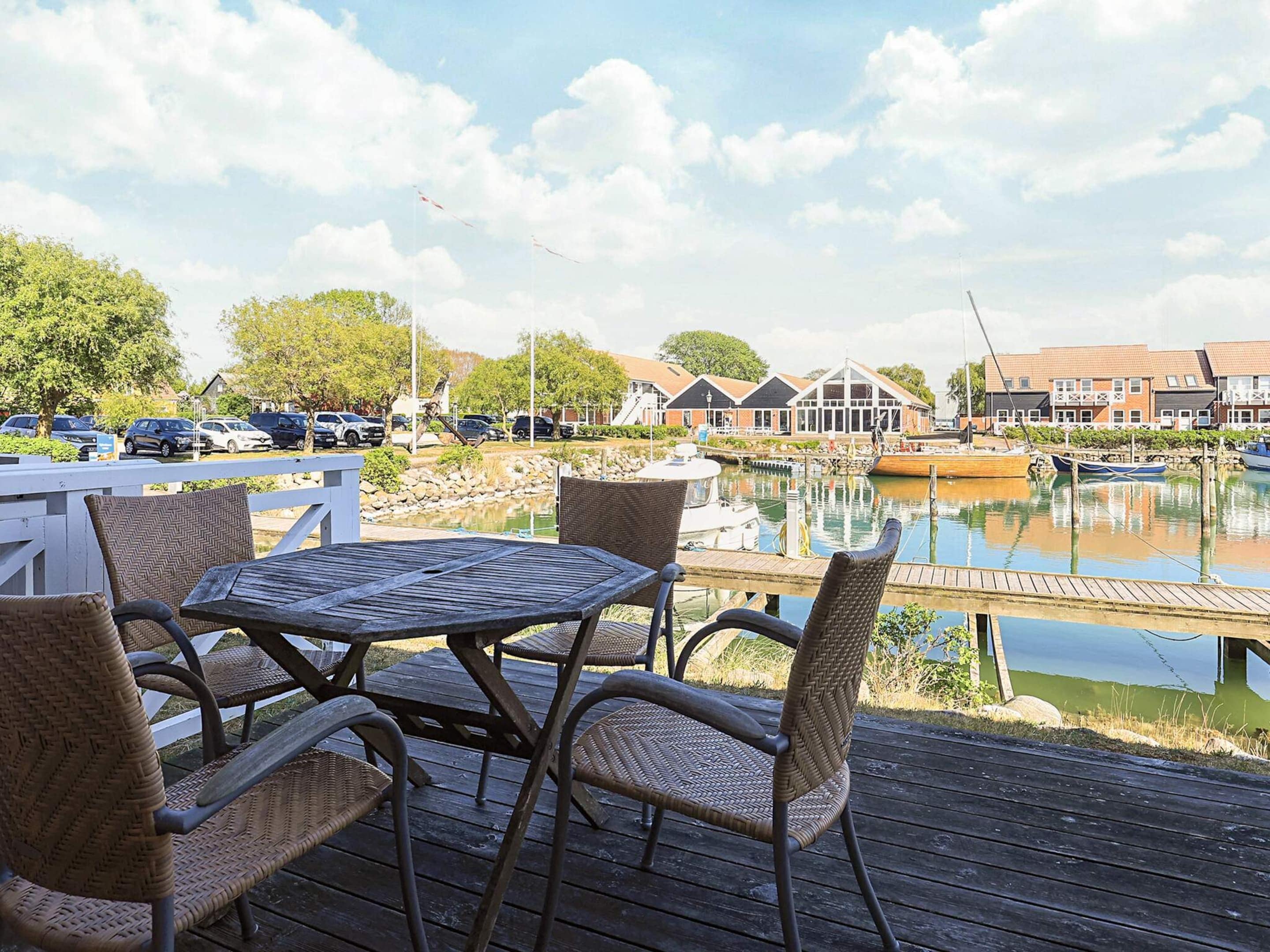 Une terrasse en bois, agrémentée de chaises et d’une table en osier, offre une vue sur une marina où l’on aperçoit des bateaux et des bâtiments en arrière-plan.
