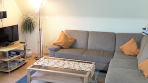 Ein Wohnzimmer mit grauem Sofa, einem Couchtisch, einem Fernseher auf einem Standfuß und einer Stehlampe.