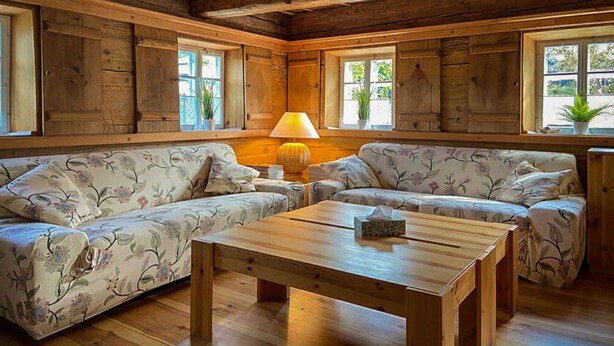 A cozy room with wooden walls and floors, two floral-patterned sofas, a wooden coffee table, and a window with a plant.