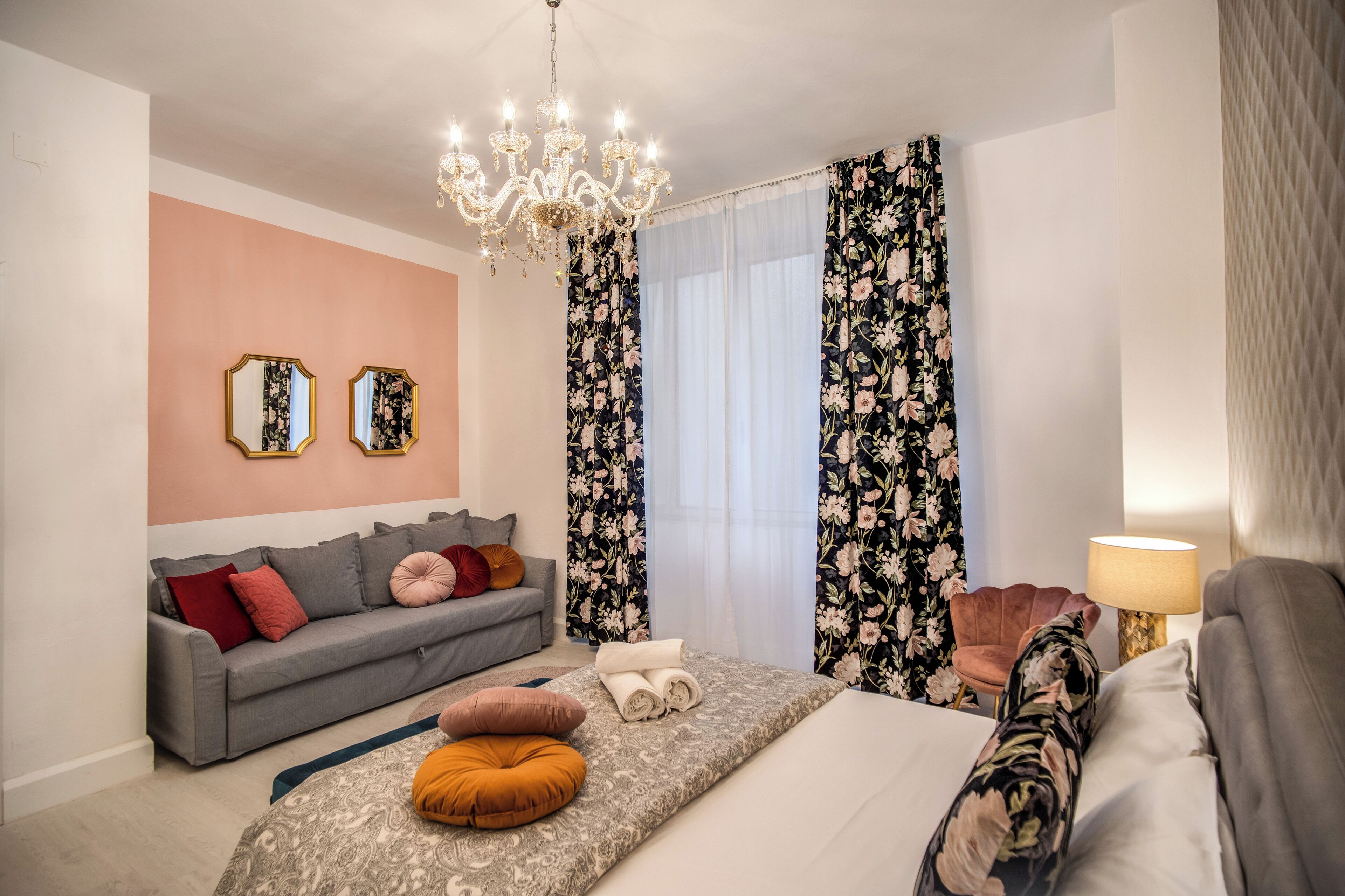 Una habitación de hotel moderna con un sofá gris, una cama con ropa de cama estampada, una araña y cortinas florales.