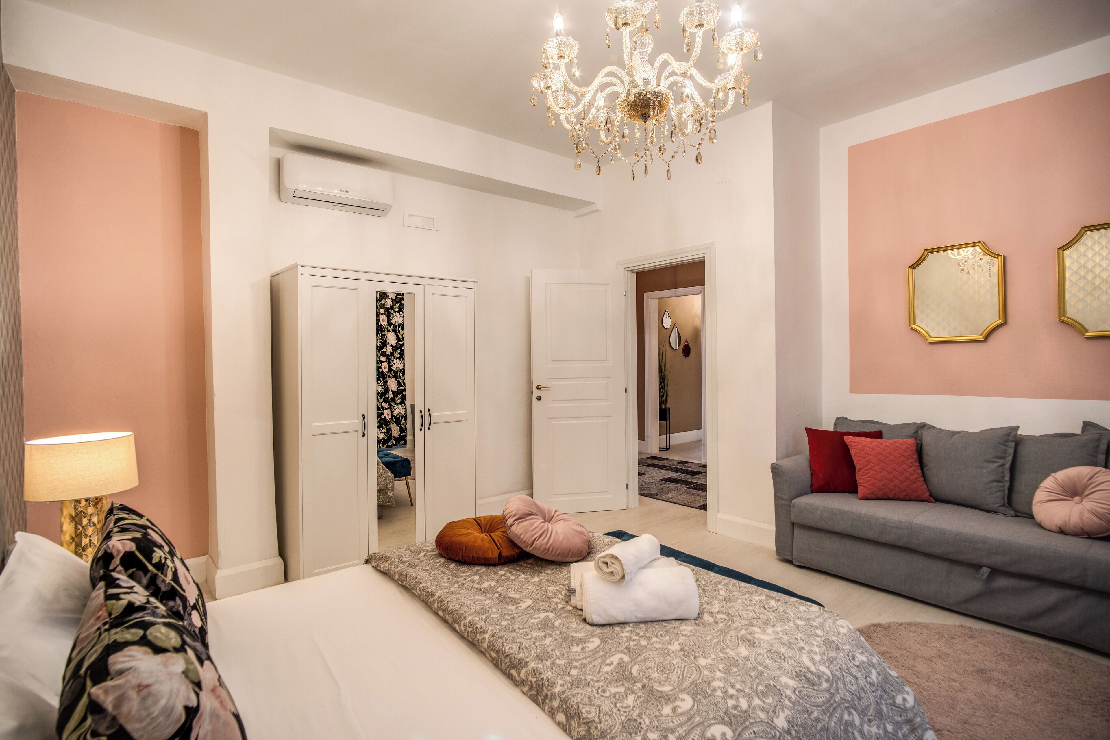 Hotellihuone, jossa on sänky, sohva, kattokruunu ja ovi, joka johtaa käytävään.