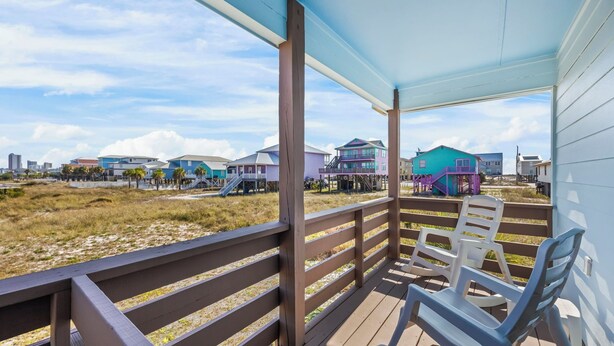 Une terrasse en bois avec des chaises en plastique blanches, donnant sur une plage bordée de maisons aux couleurs vives.