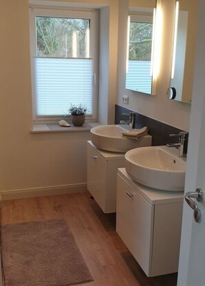 A small, clean bathroom with two sinks, a window with blinds, a potted plant, and a wooden floor. - Ferienwohnung Altes Zollhaus Burgstaaken (Fehmarn)