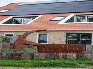Une enseigne en métal au style rustique, portant le mot « BRUG », devant une maison avec un toit en tuiles et un mur en pierre.