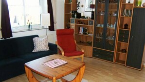 A living room with a dark blue sofa, a red armchair, and a wooden coffee table. There is a bookshelf filled with various items and a window with curtains. - "jadestrasse" 40 Merchant (Wangerland)