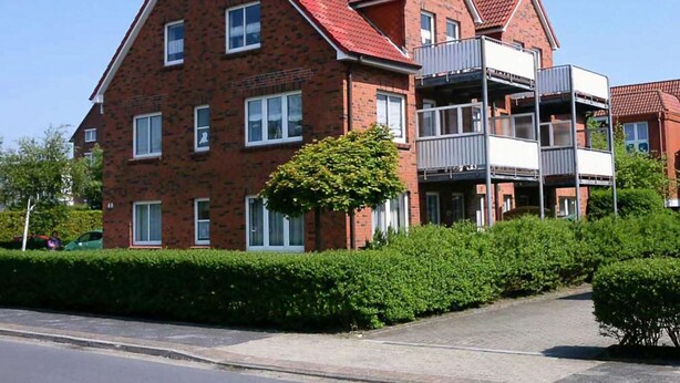 A multi-story brick building with a red-tiled roof, balconies, and a well-maintained hedge in front. - "jadestrasse" 40 Merchant (Wangerland)
