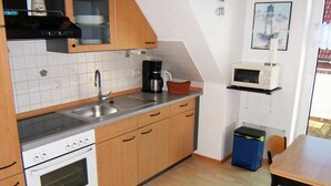 A compact kitchen with a stove, sink, and microwave. There is a dining table with chairs and a small trash bin. - "jadestrasse" 40 Merchant (Wangerland)