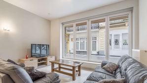 A modern living room with a sofa, a coffee table, a TV on a stand, and large windows with a view of the street. - Apartment With Modern Kitchen (Blankenberge)