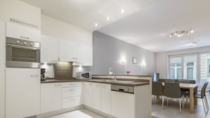 A modern kitchen with white cabinetry, a built-in microwave, and a dishwasher. There is a dining area with a table and chairs. - Apartment With Modern Kitchen (Blankenberge)