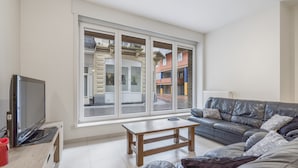 A modern living room with a large window, a sofa, a wooden coffee table, and a television on a wooden stand. - Apartment With Modern Kitchen (Blankenberge)