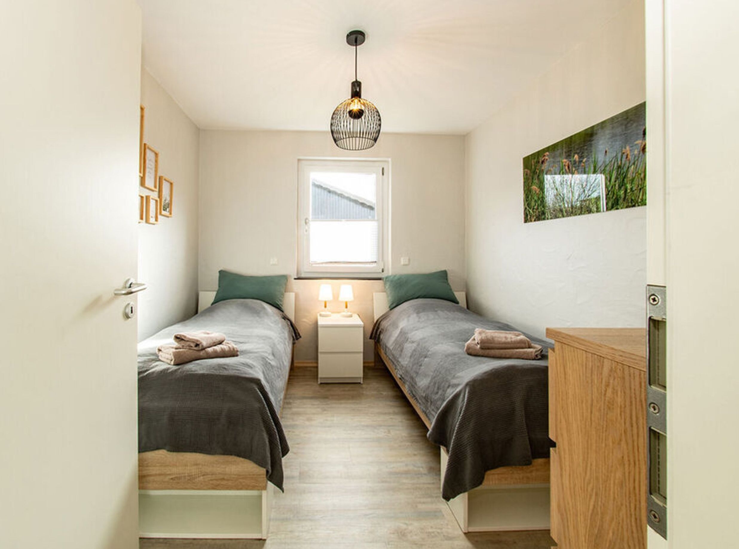 Un dormitorio pequeño y bien iluminado con dos camas, una ventana, una lámpara de noche y un cuadro en la pared.