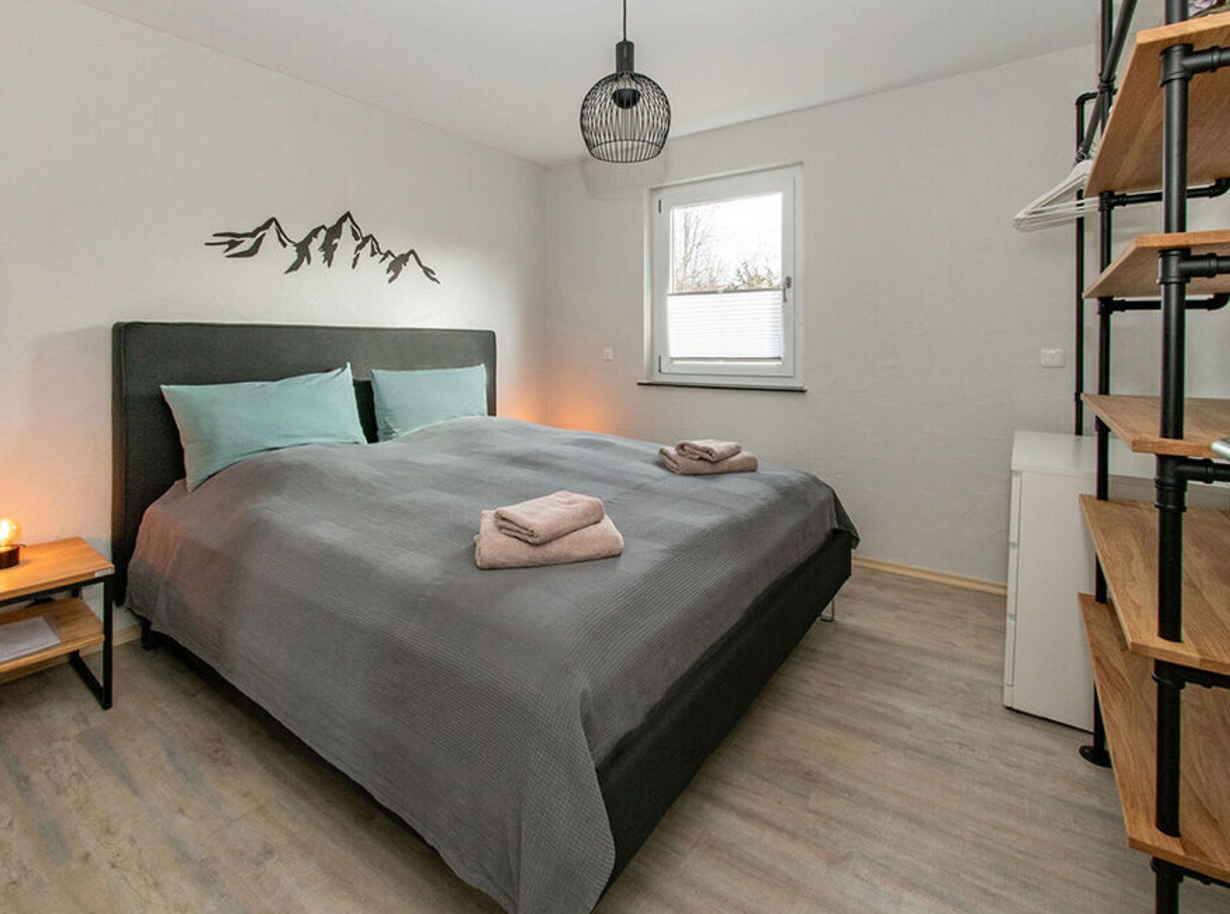 A bedroom with a bed, pillows, a bedside table, a lamp, a window, and a wall-mounted shelf.