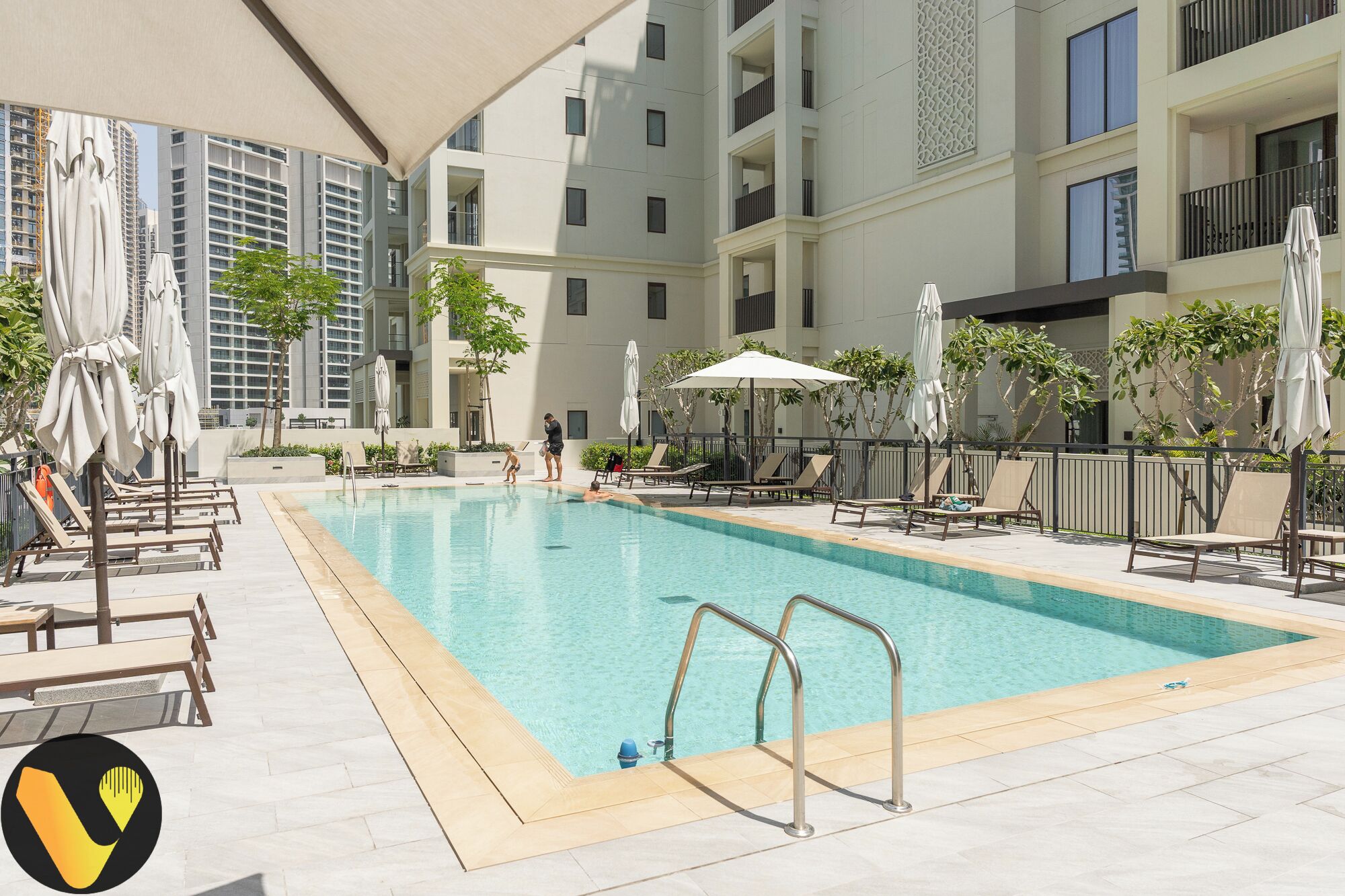 A swimming pool with lounge chairs and umbrellas, surrounded by a building with balconies and a view of other high-rise buildings.