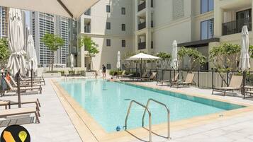 A swimming pool with lounge chairs and umbrellas, surrounded by a building with balconies and a view of other high-rise buildings.
