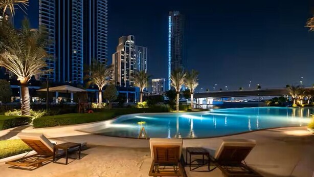 A nighttime pool area with lounge chairs, palm trees, and high-rise buildings in the background. - Vogue | Breeze Creek Beach | Waterfront Living (Dubai)