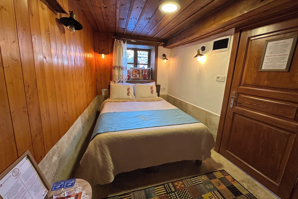 A cozy room with wooden walls, a bed with a blue bedspread, a patterned rug, and a wooden door.