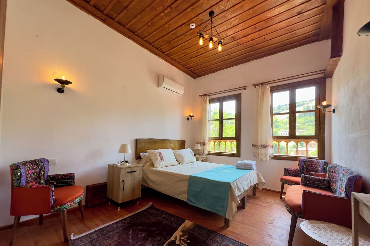 A bedroom with a wooden ceiling, a bed with a blue bedspread, two nightstands, a chair, a table, and a window with curtains.