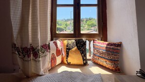 A window with a view of a hilly landscape, a bed with a white blanket, and a curtain with floral patterns. - Çirkince Konak Şirince (Selçuk)