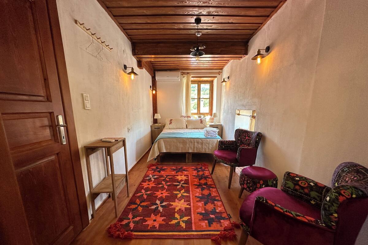 A narrow hallway leading to a bedroom with a bed, a chair, a table, and a patterned rug.