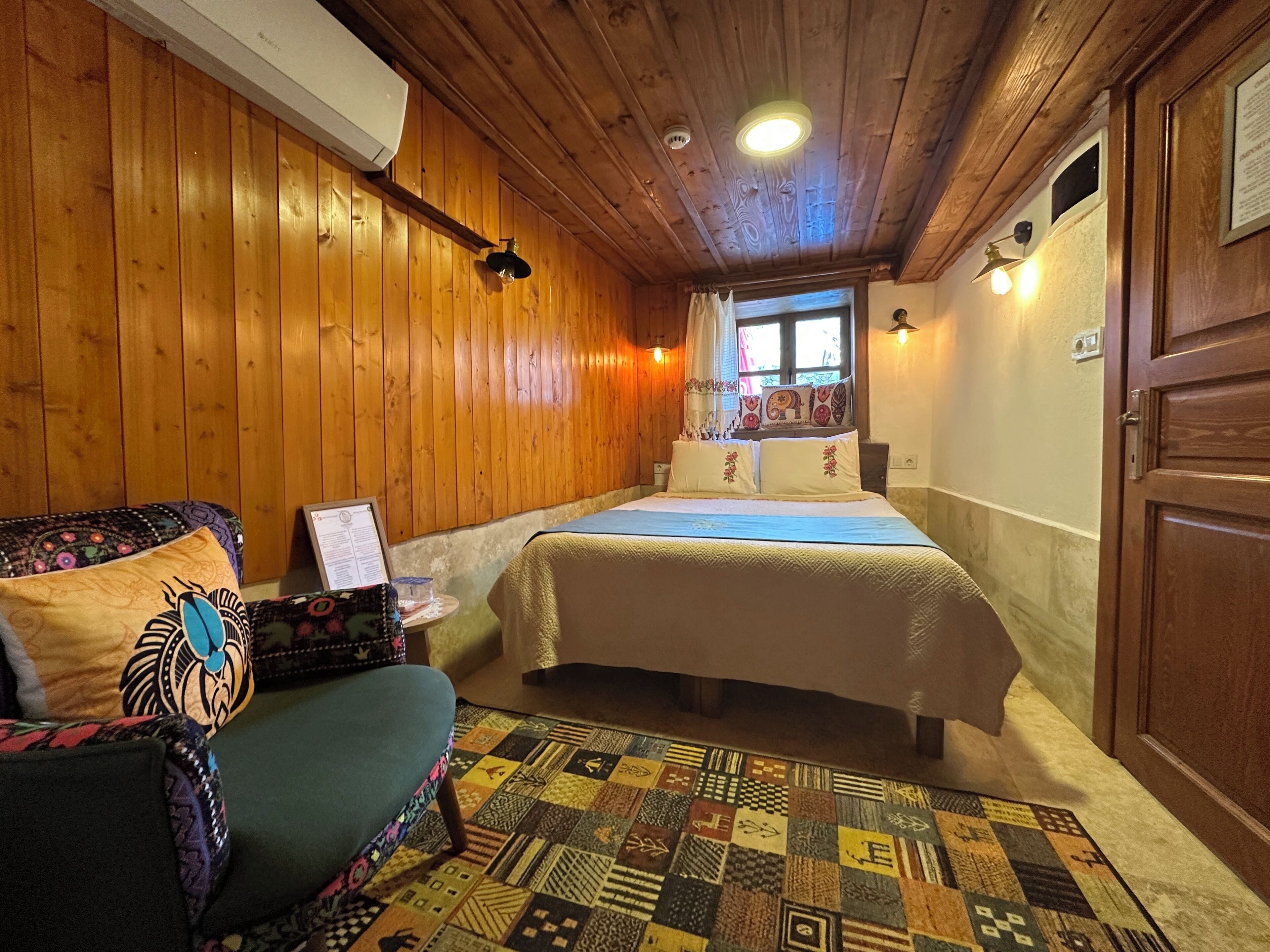 A cozy room with wooden walls, a bed with patterned bedding, a small table with a framed picture, and a colorful quilt on a chair.
